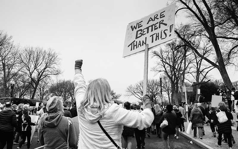 Within a protest a women holds up a sign saying, “We are better than this!”
