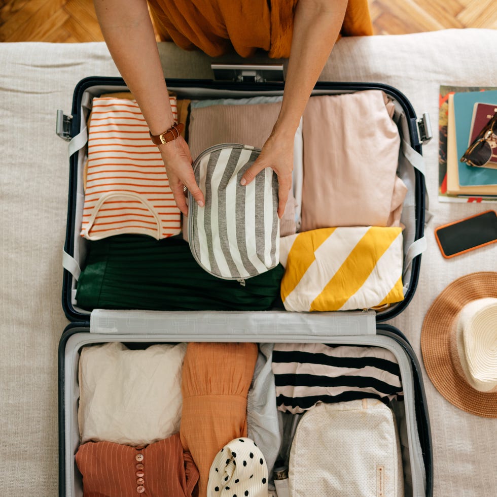 Woman Putting a Cosmetic Bag in Her Suitcase shot from above of an anonymous woman packing things in her suitcase on the bed she is holding and putting a grey and white stripped cosmetics bag in there is her mobile phone, books and sunglasses on the bed