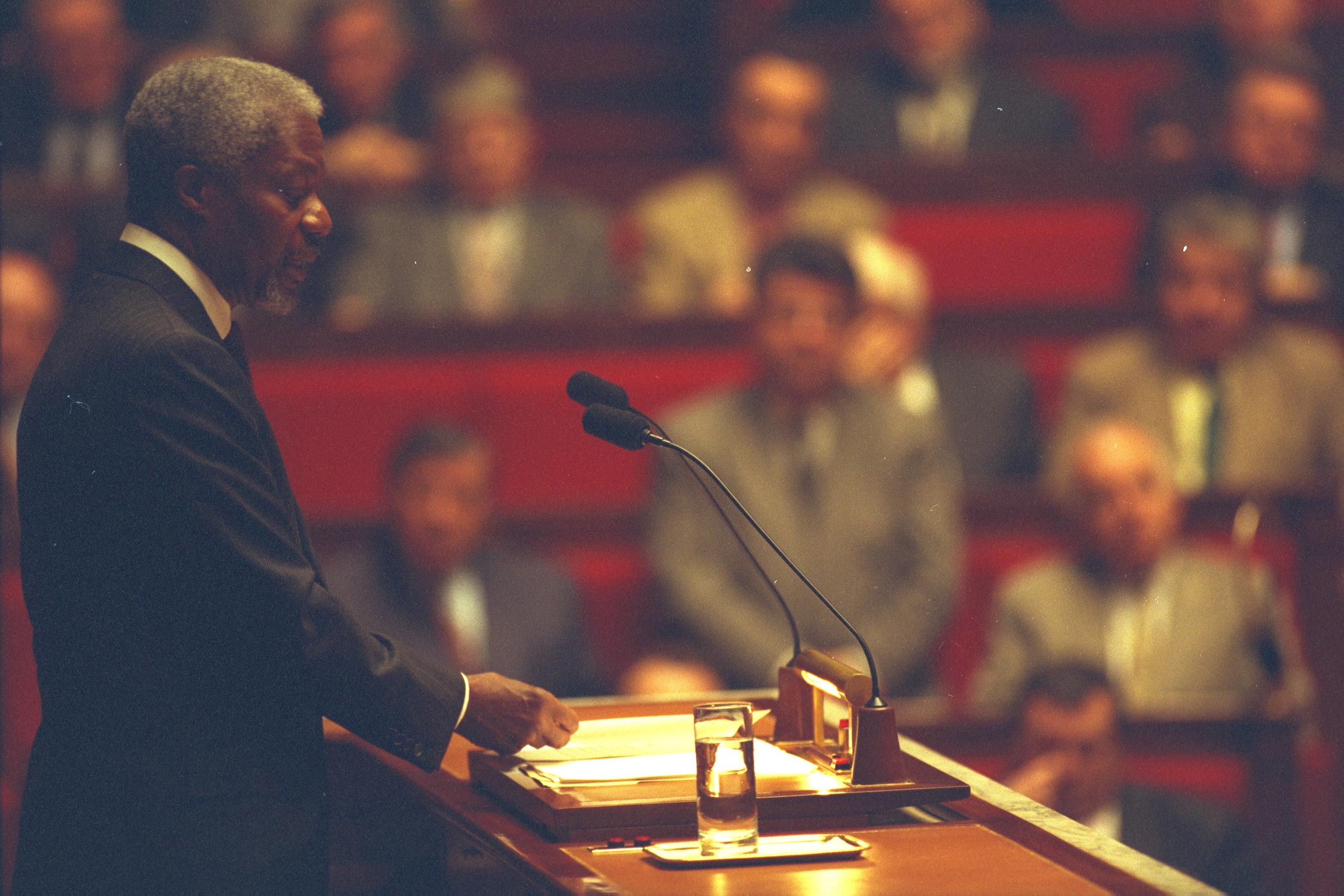 Kofi Annan speaks at a lectern with two microphones before a seated audience. The journalistic, action-oriented photo captures him in profile, emphasizing his role as a global leader in a formal, high-stakes setting.